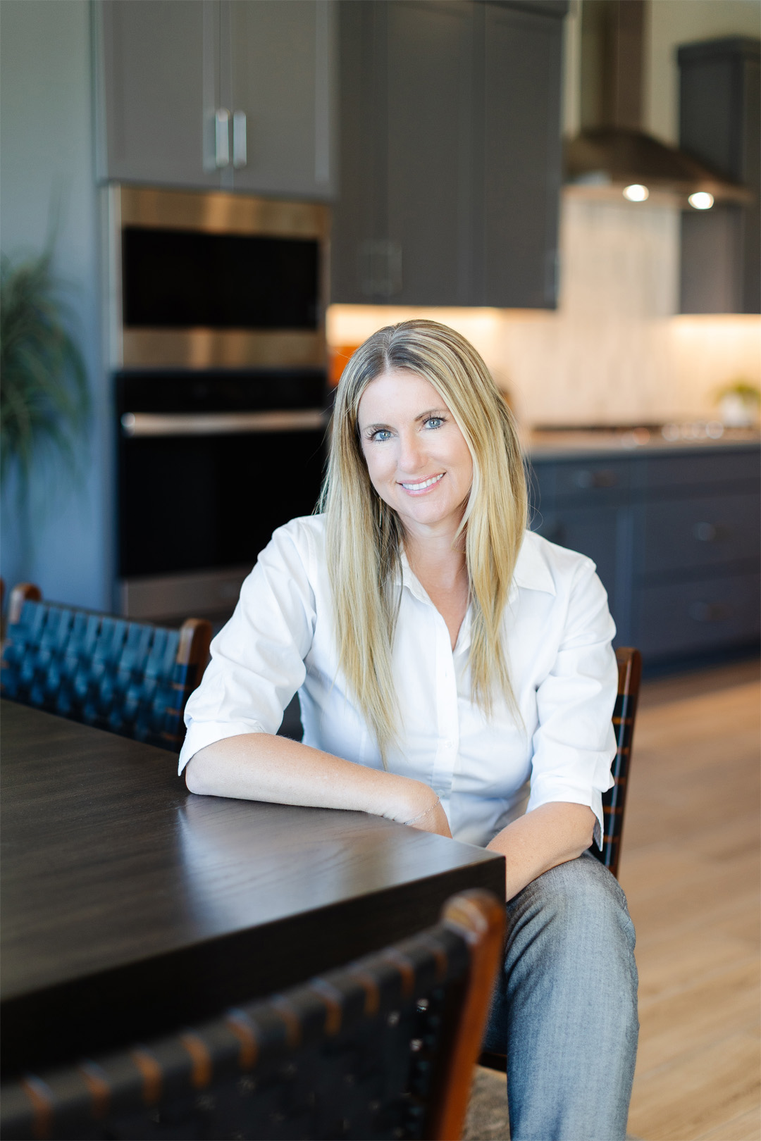 sarahluzi Smiling woman with long blonde hair, wearing a white blouse, seated at a modern dining table in a stylish kitchen.